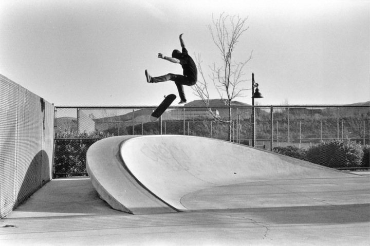 A skateboarder is captured mid-air above a concrete skate park ramp with a fence and hills in the background.