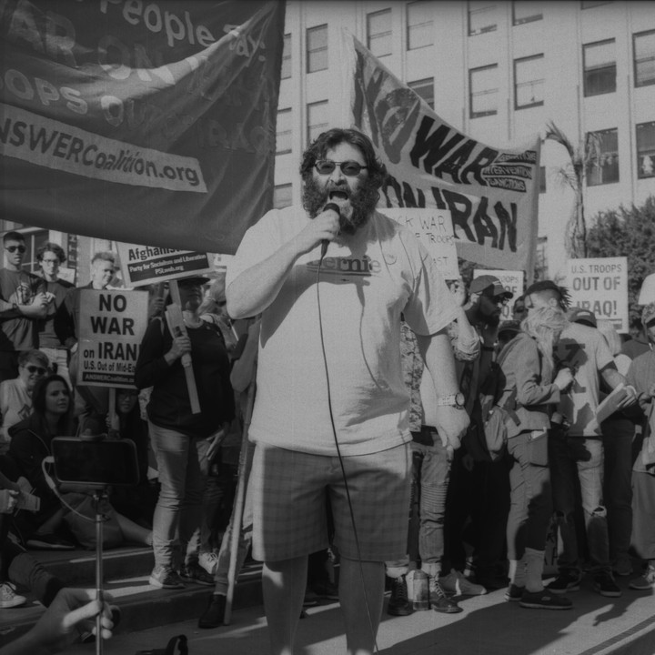 A man with a beard and sunglasses speaks into a microphone at a protest with signs that say "No War on Iran" and "U.S. Troops Out of Iraq."