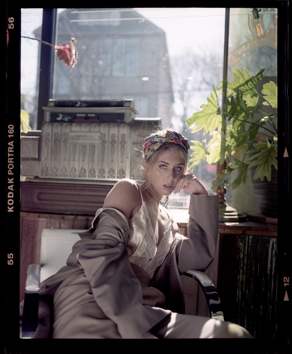 A woman wearing a headscarf and a coat sits in a chair next to a window with a plant and a cash register.