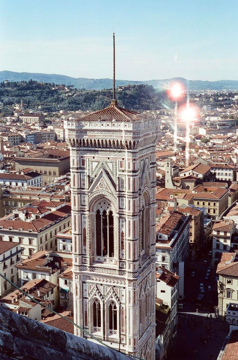 A tall, ornate bell tower rises above a city with red-tiled roofs and distant hills under a bright sky with lens flare.