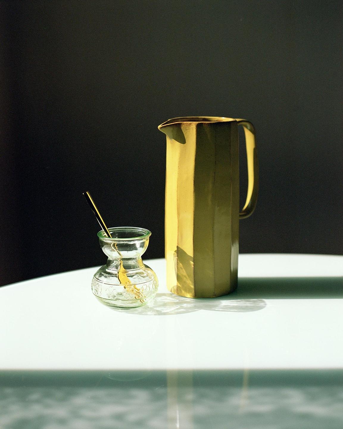 A yellow pitcher and a small glass vase with a stirrer are arranged on a white surface.