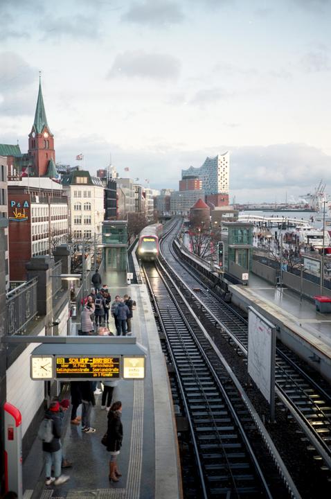 A train approaches a station platform where people are waiting, with a city skyline and harbor in the background.
