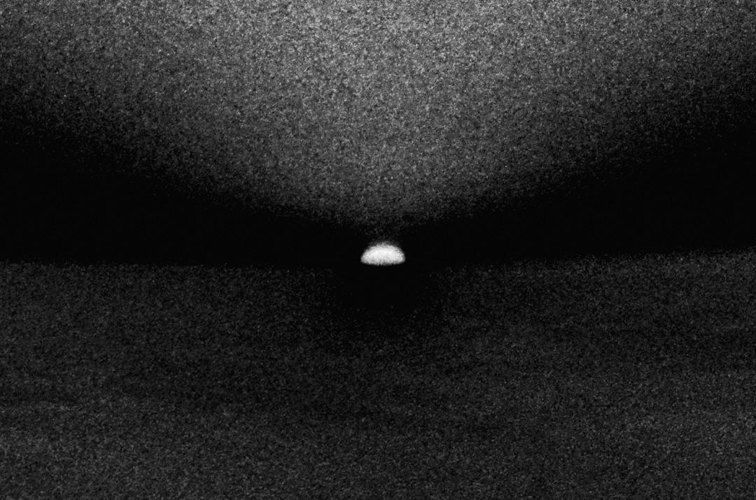 A small, bright, half-circle shape is visible on the horizon in a grainy, black and white landscape.