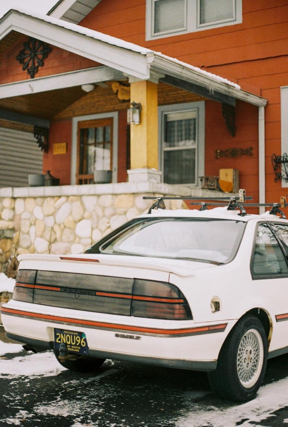 A white car with a roof rack is parked in front of an orange house with snow on the ground.