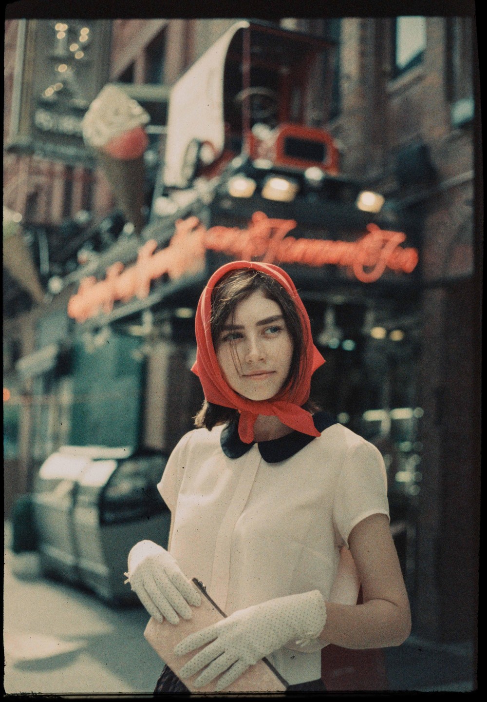 A woman wearing a red headscarf and white gloves stands on a street with a neon sign in the background.