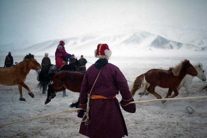 A person in traditional clothing stands in a snowy landscape with horses and other people in the background.