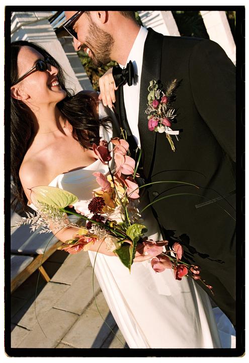 A couple is smiling at each other, the woman holding a bouquet of flowers.