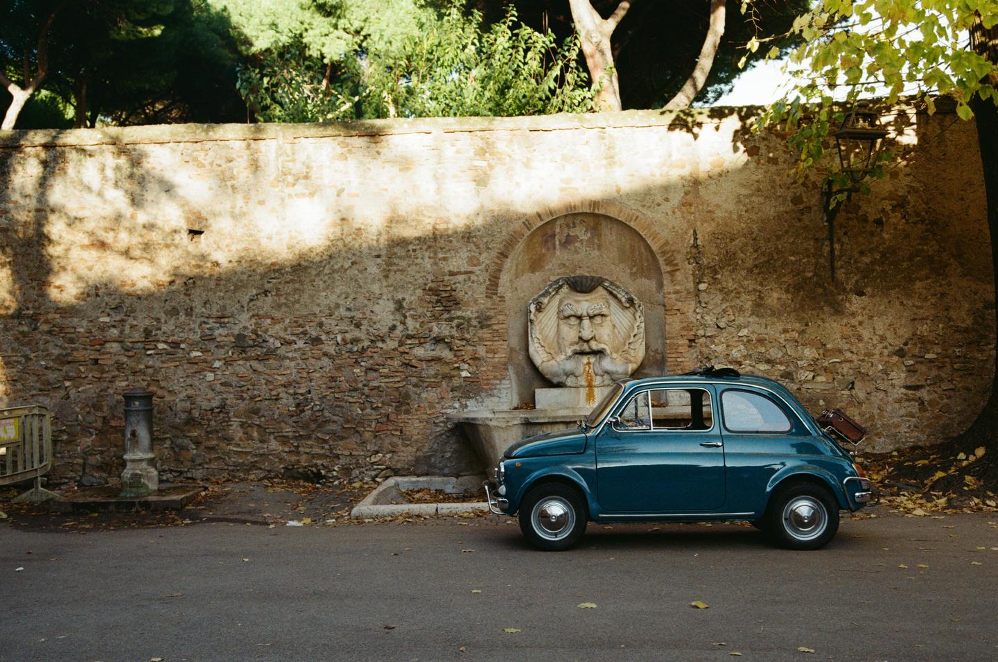 A blue car is parked next to a stone wall with a decorative fountain.