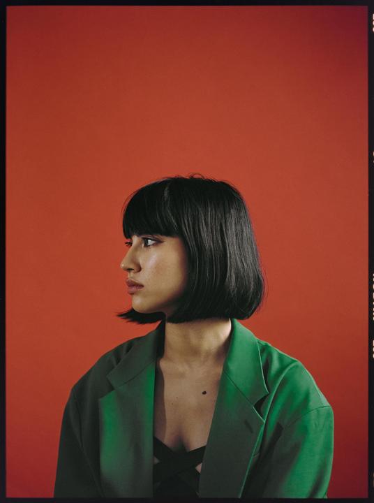 A woman with short dark hair and a green jacket looks to the left against a red background.