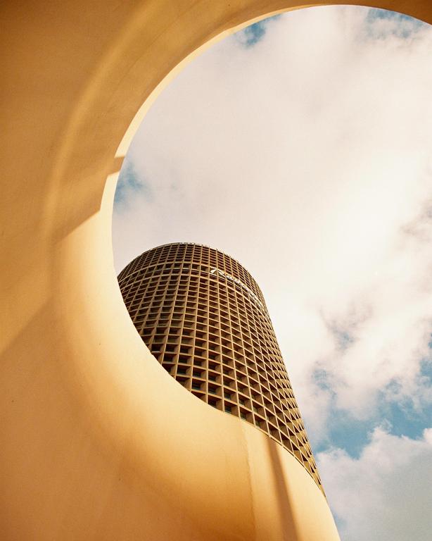 A tall, cylindrical building with a grid pattern on its facade is framed by a curved, light-colored structure against a cloudy sky.