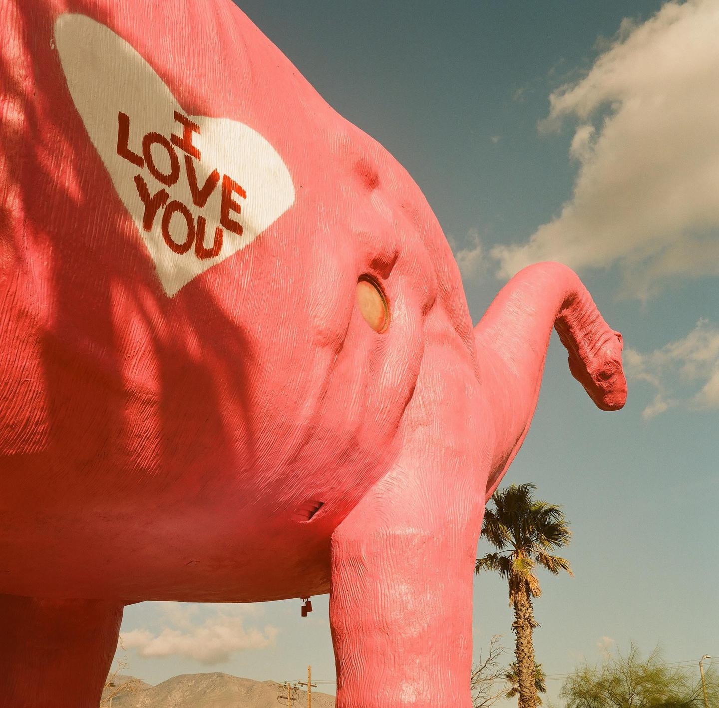 A large pink dinosaur statue with "I LOVE YOU" written on its side stands under a cloudy sky.
