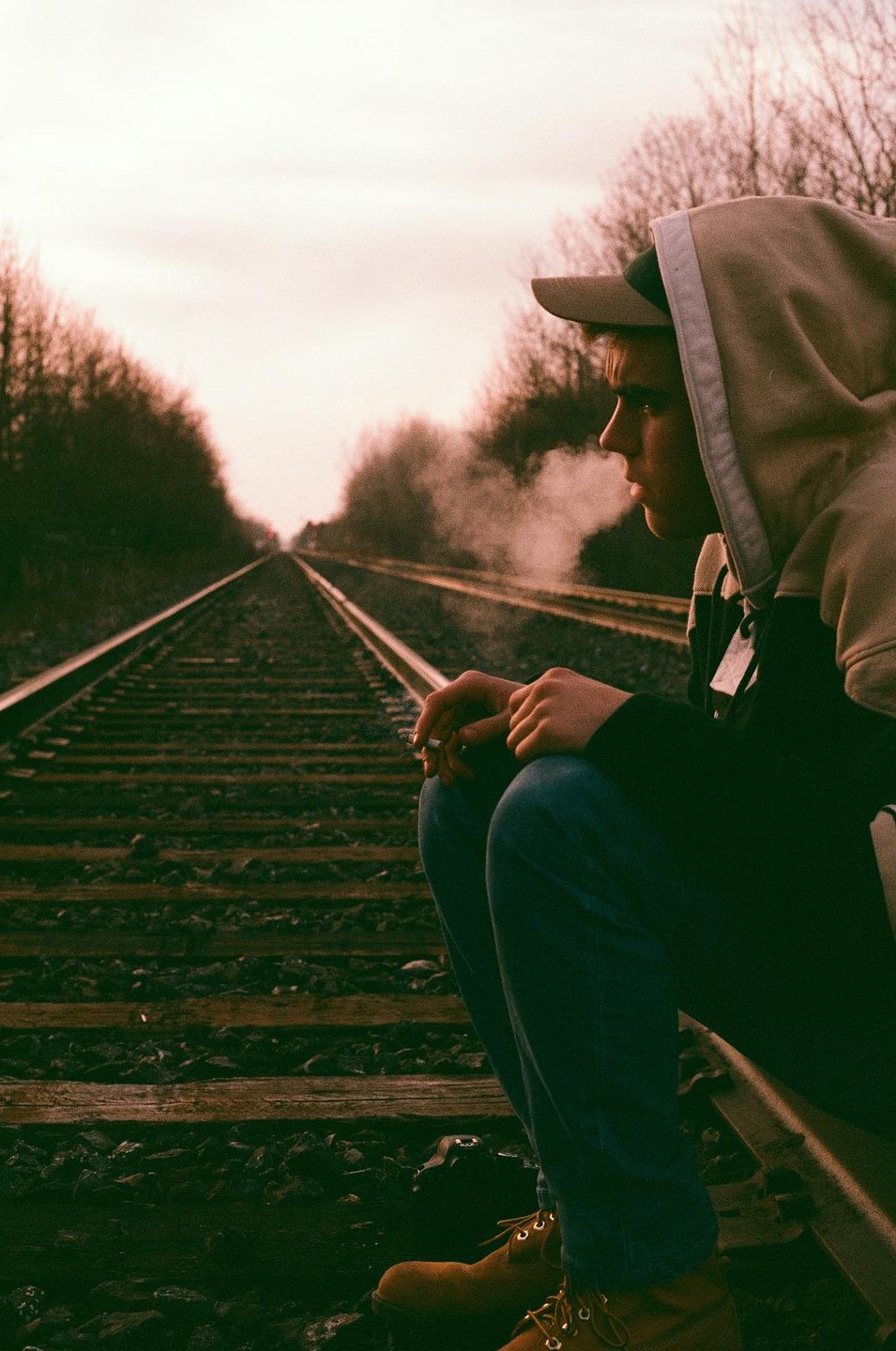 A young man in a hoodie and baseball cap crouches on railroad tracks, holding a cigarette with smoke exhaling from his mouth.