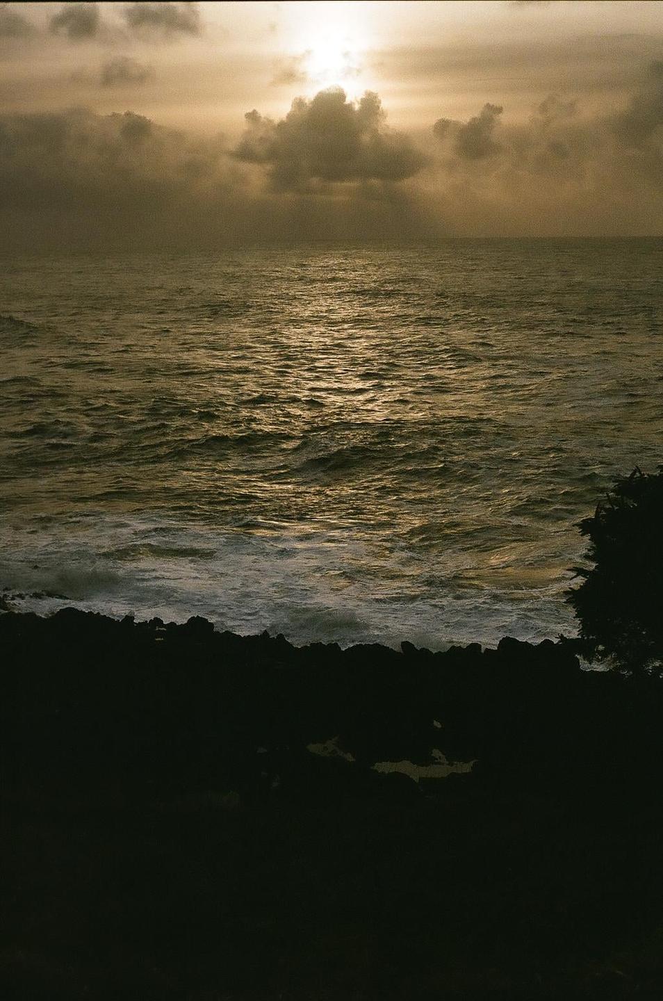 The sun shines through clouds over a rocky coastline and ocean waves.