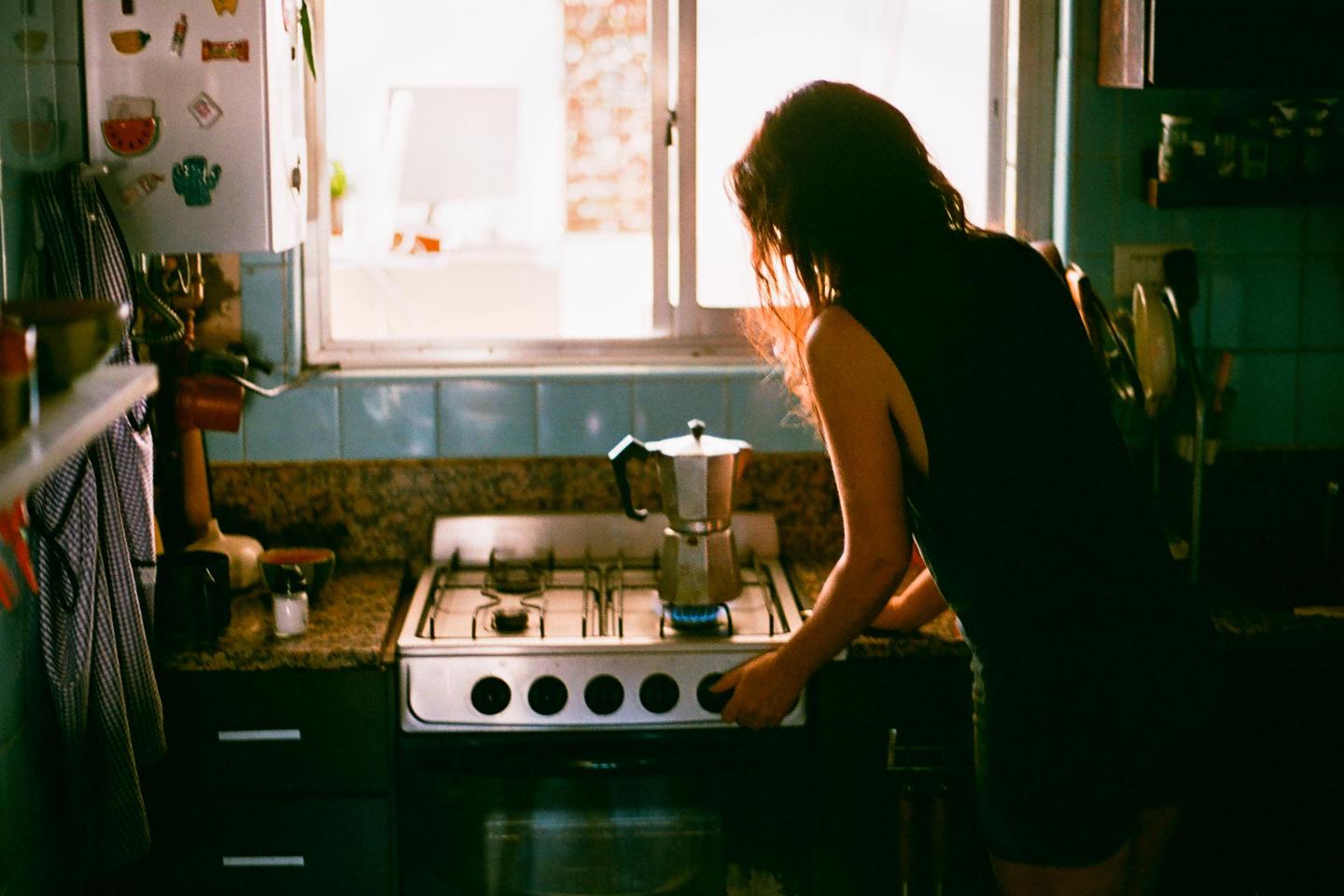 A woman stands in a kitchen, turning on a gas stove with a moka pot on it.