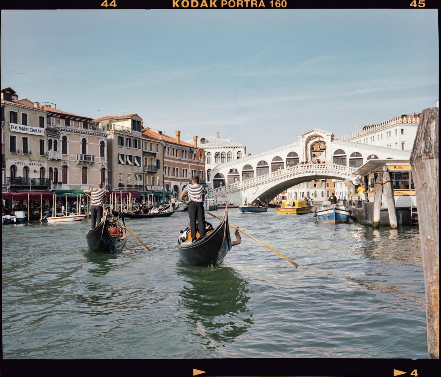 Two gondoliers in striped shirts steer their gondolas on a canal with the Rialto Bridge and buildings in the background.
