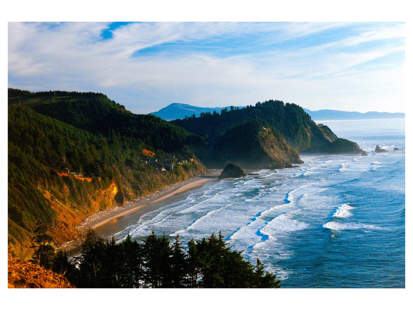 A long view of a coastline with a beach, ocean waves, and forested cliffs under a blue sky.