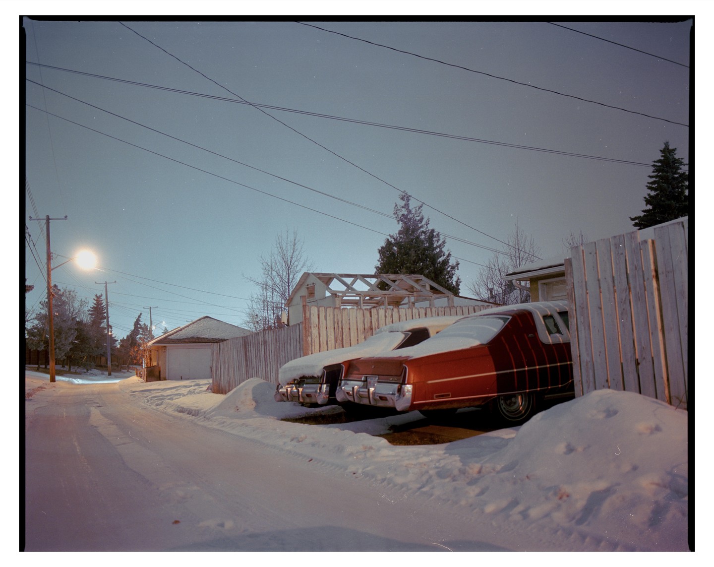 Two vintage cars are parked next to a wooden fence on a snow-covered street at night, illuminated by a streetlamp.