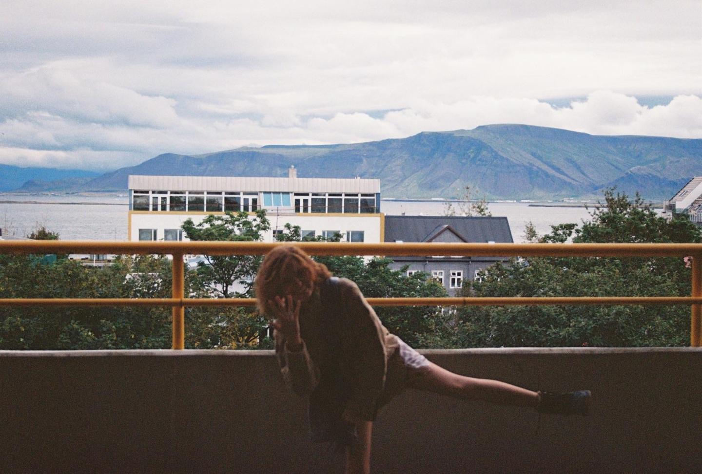 A woman poses on a balcony overlooking a body of water and mountains under a cloudy sky.