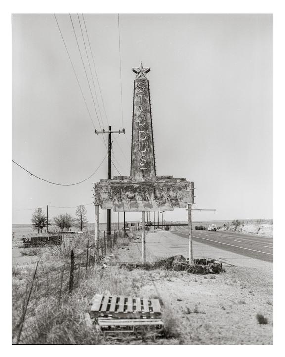 A tall, weathered sign for "Stardust" with a star on top stands beside a road in a dry, rural landscape.