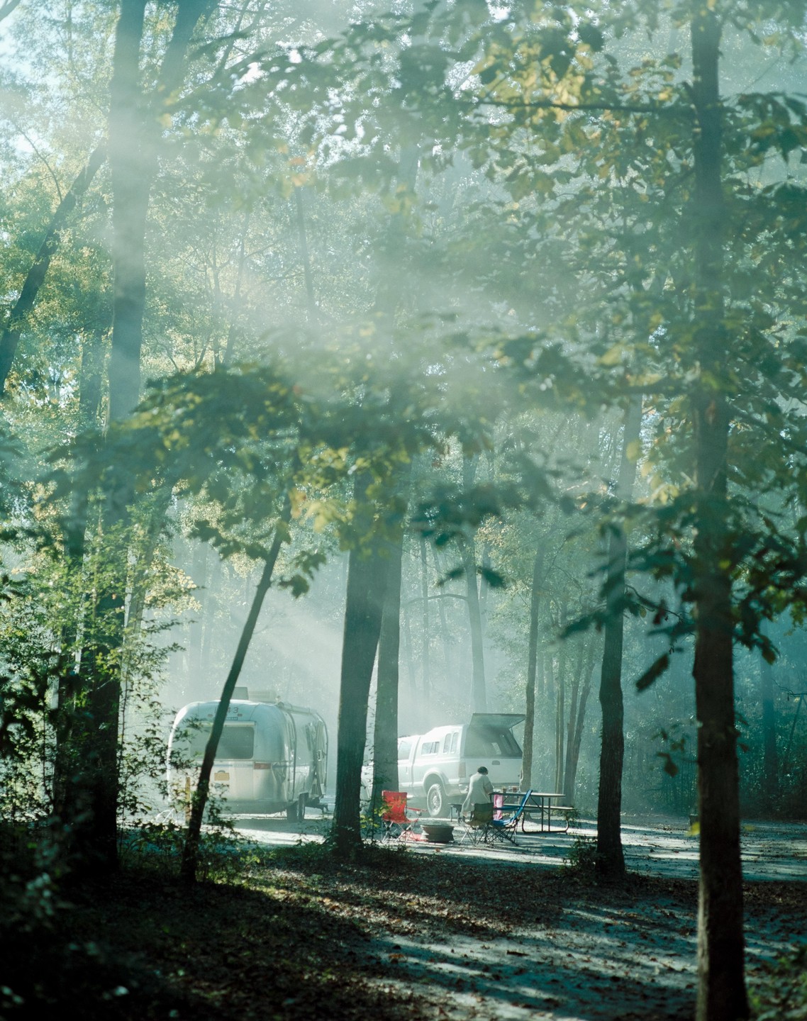 A person stands by a truck and a travel trailer in a hazy forest.