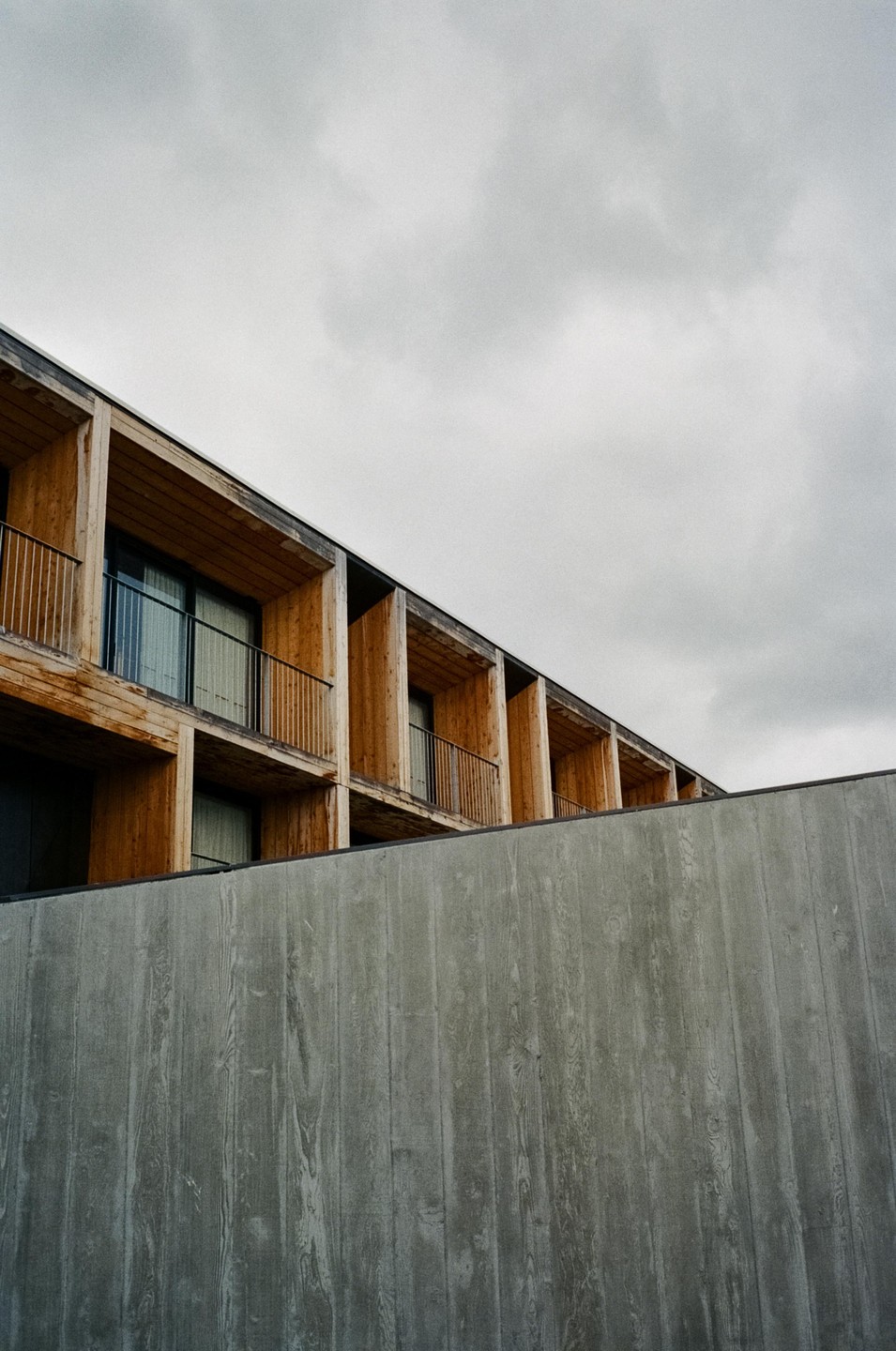 A modern building with wooden balconies and a concrete wall in the foreground is shown under a cloudy sky.