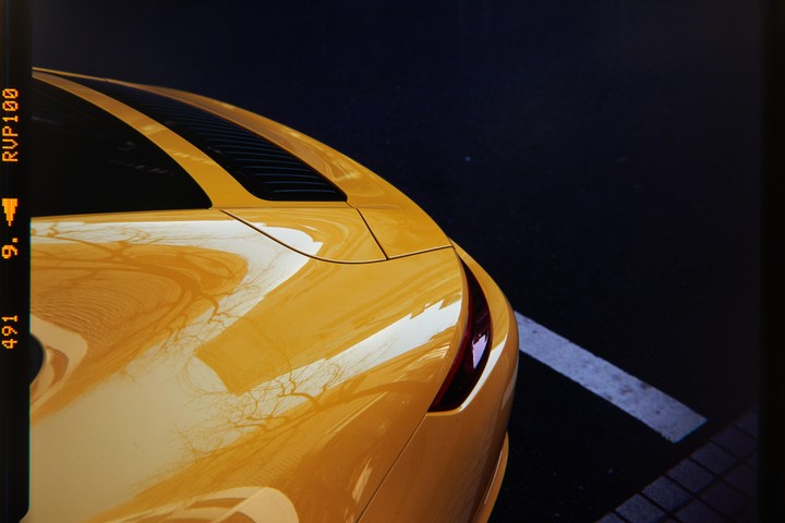 The rear of a yellow car is parked on a dark surface with a white line.
