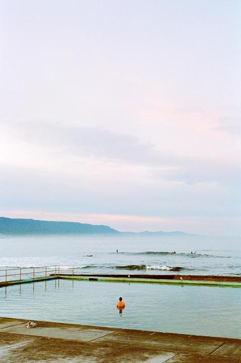 A person in an orange shirt stands in a concrete ocean pool with mountains and surfers in the background.