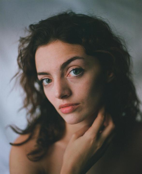 A woman with dark, curly hair and green eyes looks directly at the viewer, with one hand touching her neck.