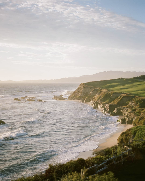 Waves crash against a sandy beach and rocky cliffs with green grass on top under a hazy sky.