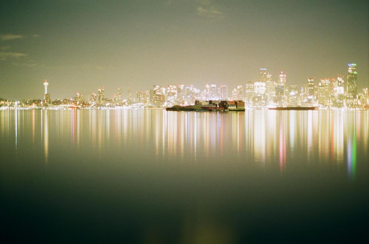 A city skyline with a prominent tower is reflected in the calm water at night, with a barge visible in the middle ground.