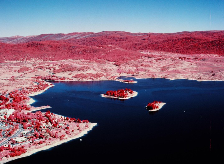 An aerial landscape shows a lake with two small islands, surrounded by hills and a town with roads, all rendered in infrared colors with deep blue water and bright red foliage.