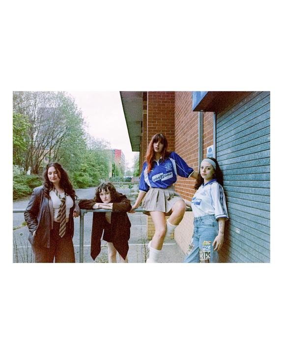 Four women pose for a group portrait on a street next to a brick building with a metal gate.