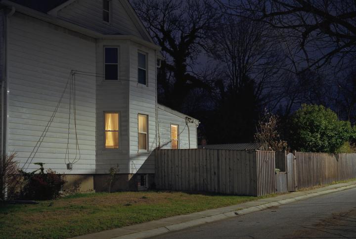 A white house with illuminated windows stands beside a wooden fence and a street at night.
