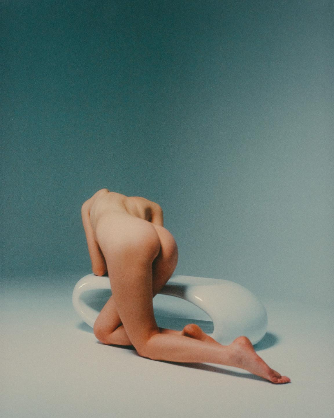 A nude woman kneels on a white surface, leaning on a white, curved object, with her back to the viewer.