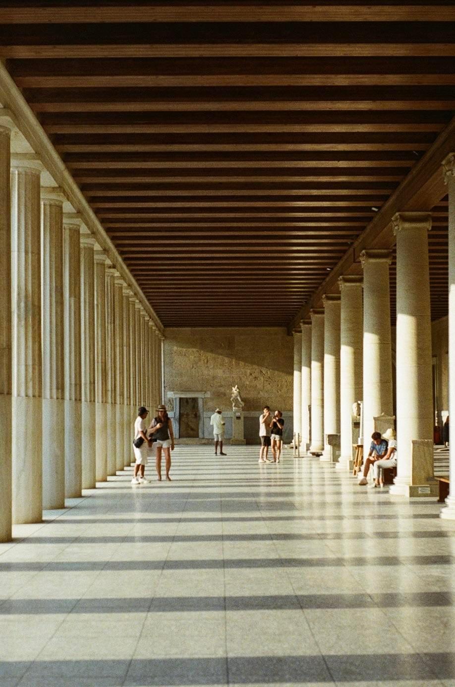 A long colonnade with many columns and a wooden ceiling is filled with people and shadows.