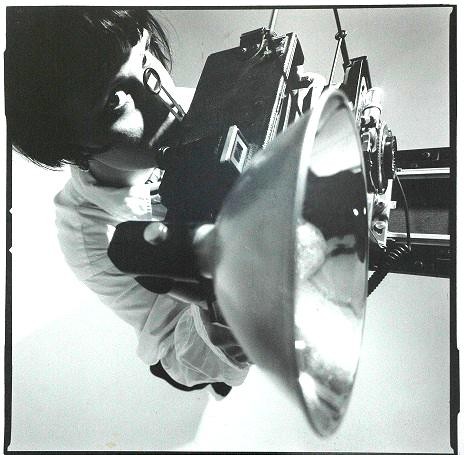A woman looks through the viewfinder of a large format camera with a flash attached.