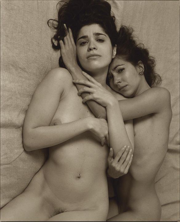 Two women lie together, one embracing the other, both looking towards the viewer.