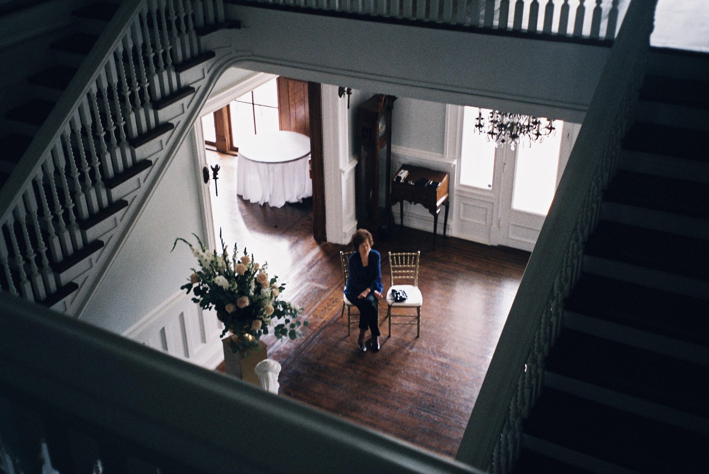 A woman sits on a chair in a large hall with a grand staircase and a floral arrangement.