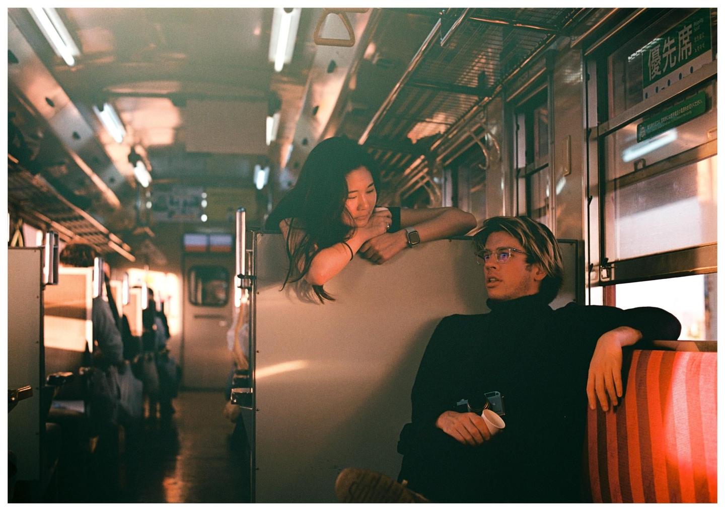 A woman leans over a partition to talk to a man sitting on a train.