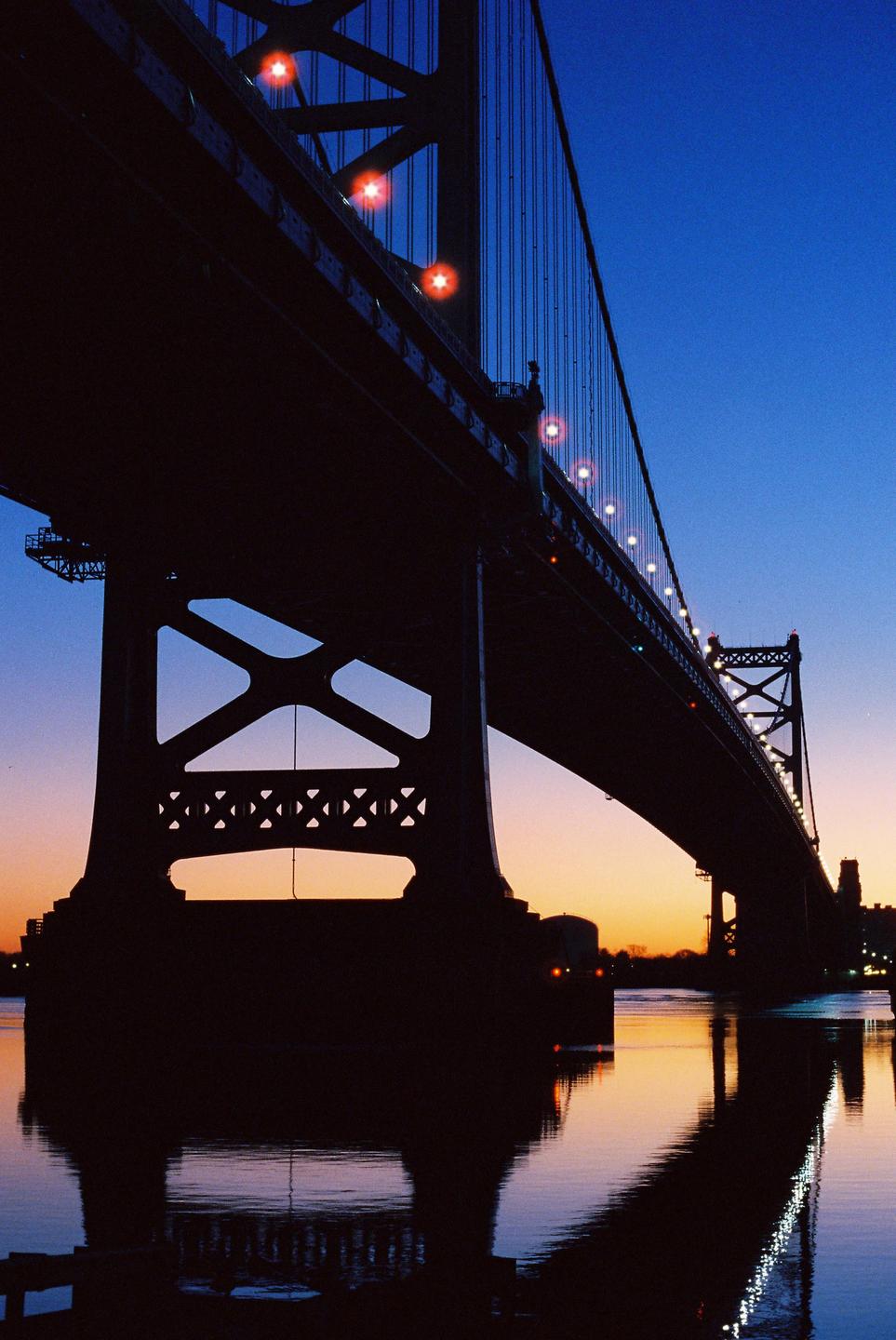 A large bridge is silhouetted against a colorful sky at dusk, with its reflection visible in the water below.