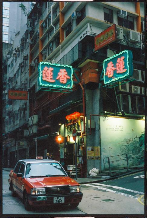 A red taxi is parked on a street with neon signs and tall buildings in the background.