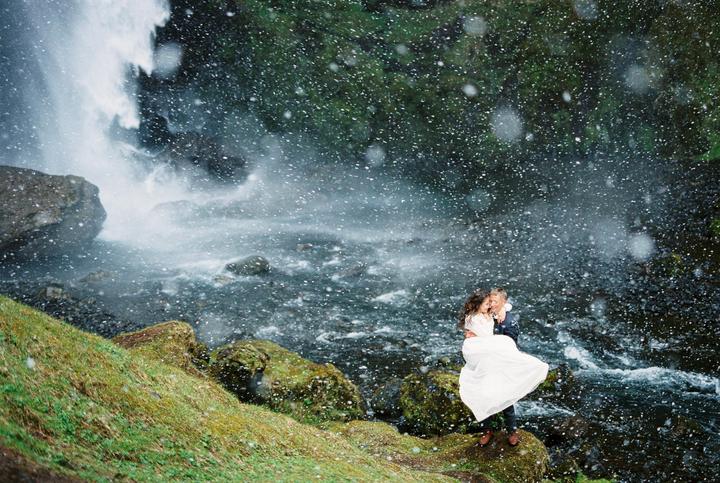 A couple embraces on a mossy rock next to a waterfall with water droplets in the air.