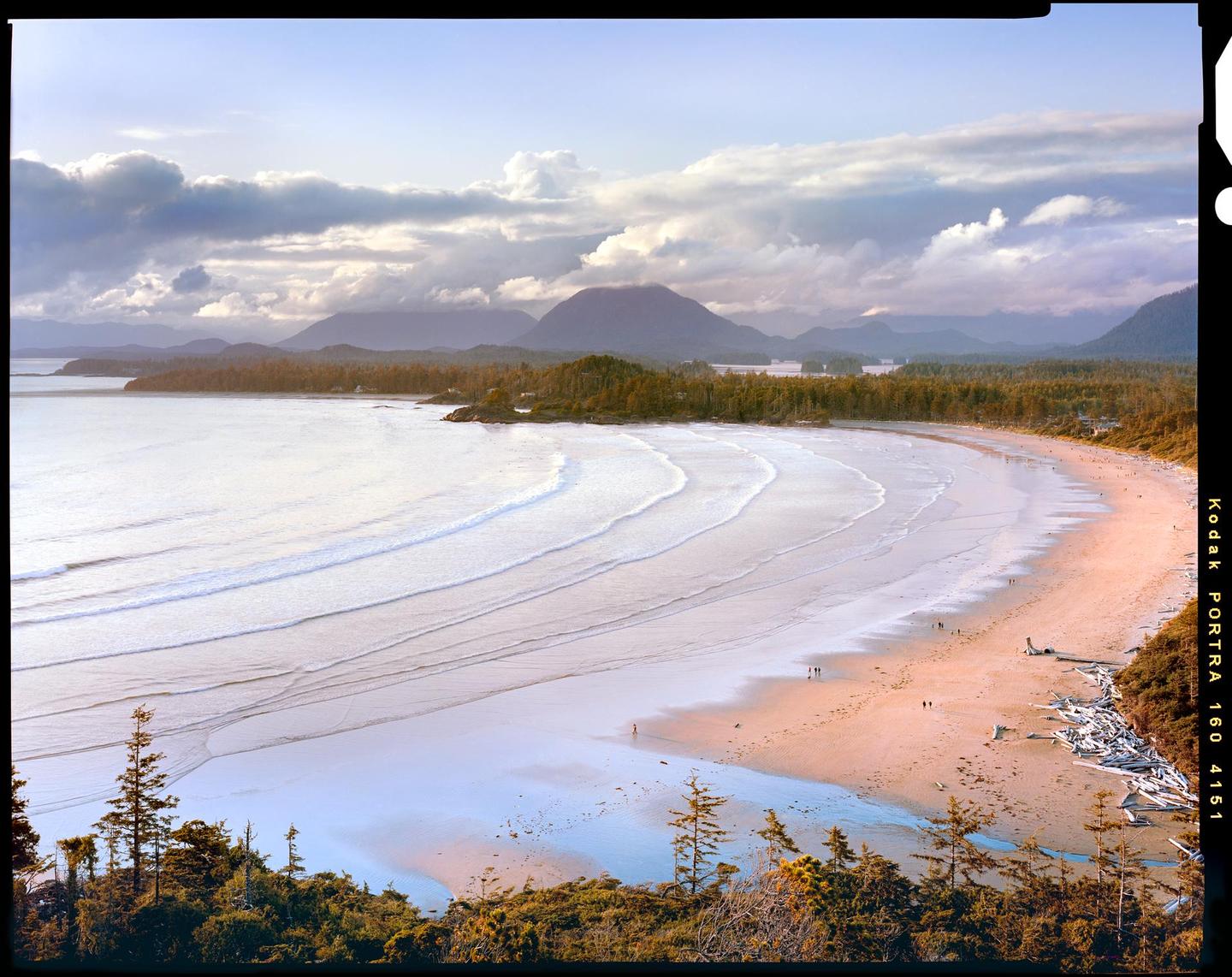 A wide sandy beach with gentle waves is visible from above, with a forest and mountains in the distance under a cloudy sky.