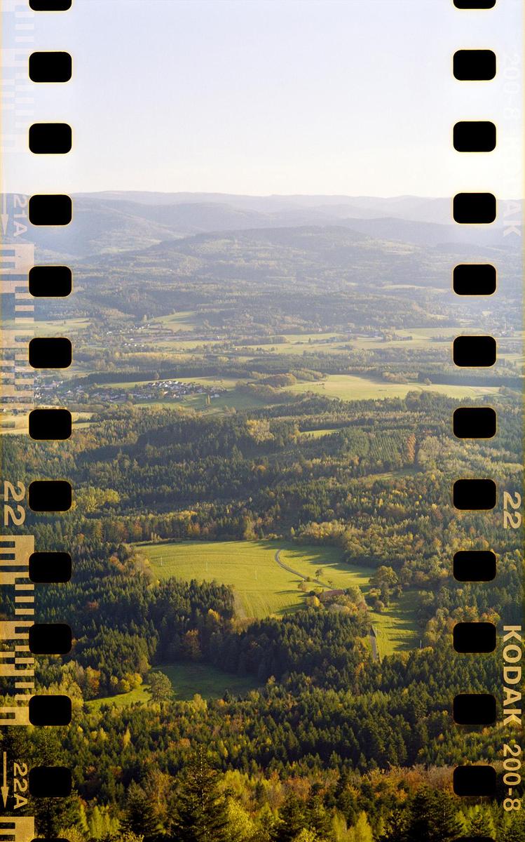 An aerial view shows a vast landscape of rolling hills, forests, and fields under a bright sky.