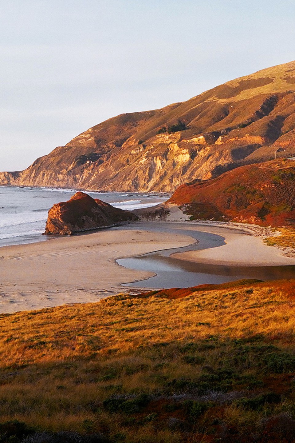 A river winds through a sandy beach towards the ocean, with hills rising in the background.