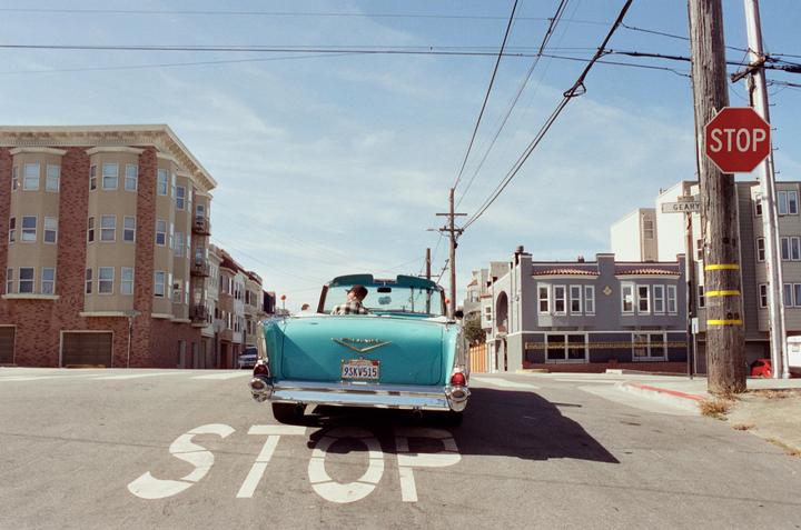 A turquoise convertible with a man driving is stopped at an intersection with a stop sign and buildings on either side.