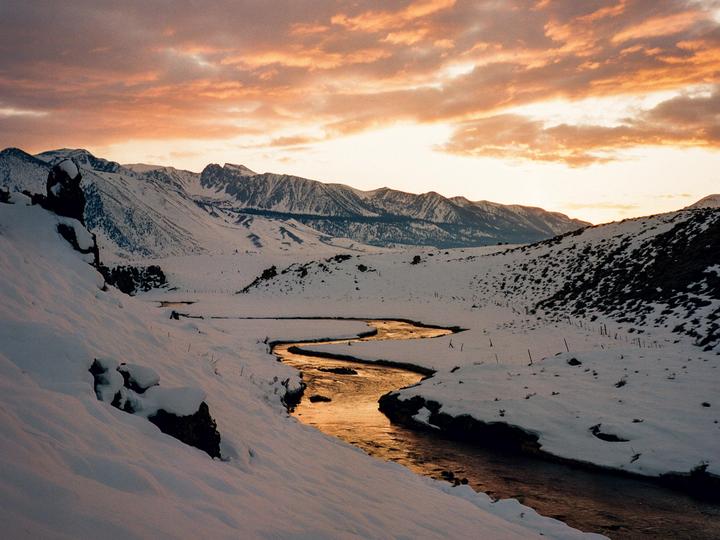 A winding river reflects the orange sky as it flows through a snow-covered mountain landscape.