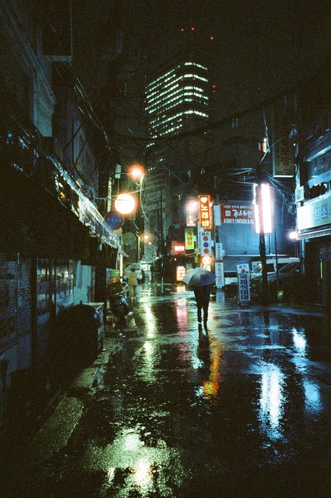 A person walks with an umbrella down a wet street at night, reflecting neon signs and city lights.