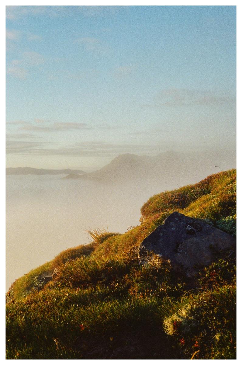 A rocky, grassy hillside overlooks a misty valley with mountains in the distance.
