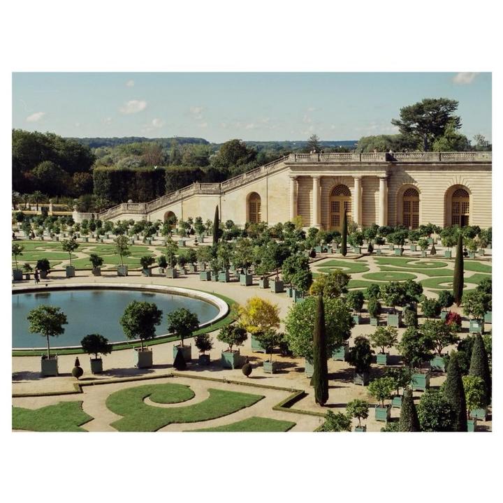 A large formal garden with a circular pond and a grand building with columns and arched windows in the background.